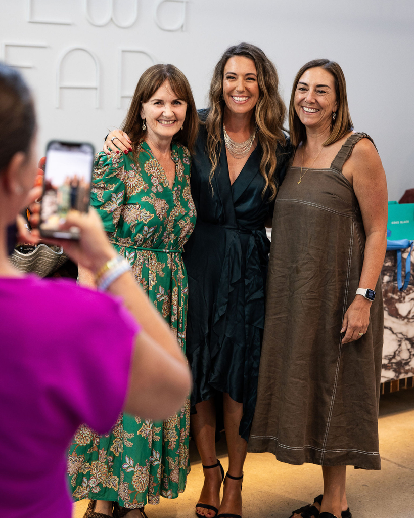 Three women posing for a photo with a fourth person taking the picture, indoors.