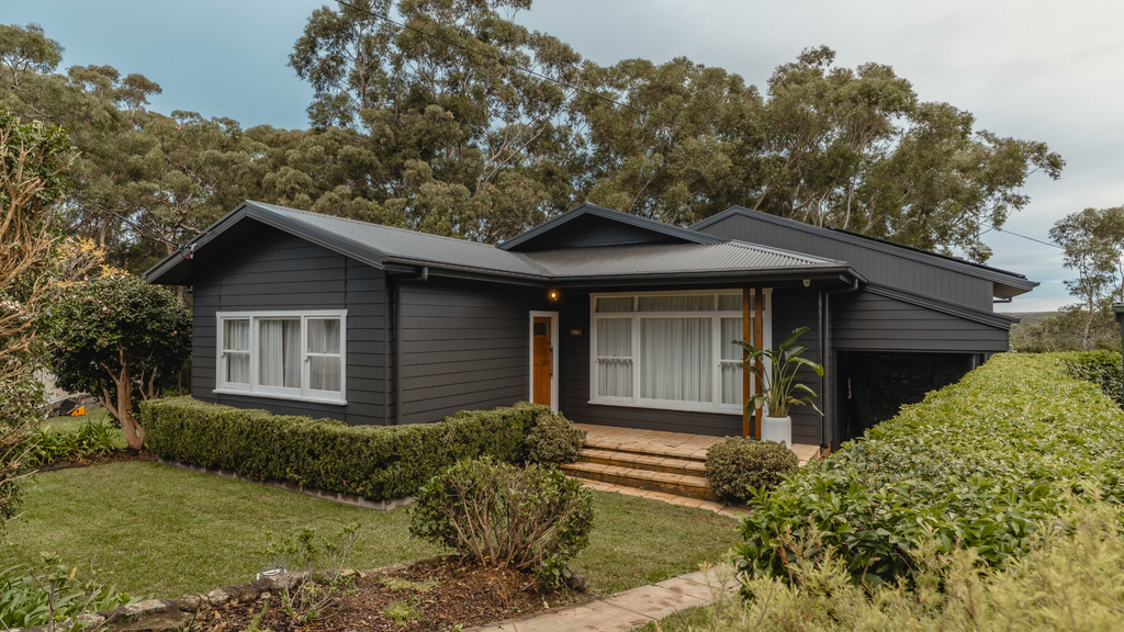 Modern house with dark exterior and white windows, surrounded by greenery.